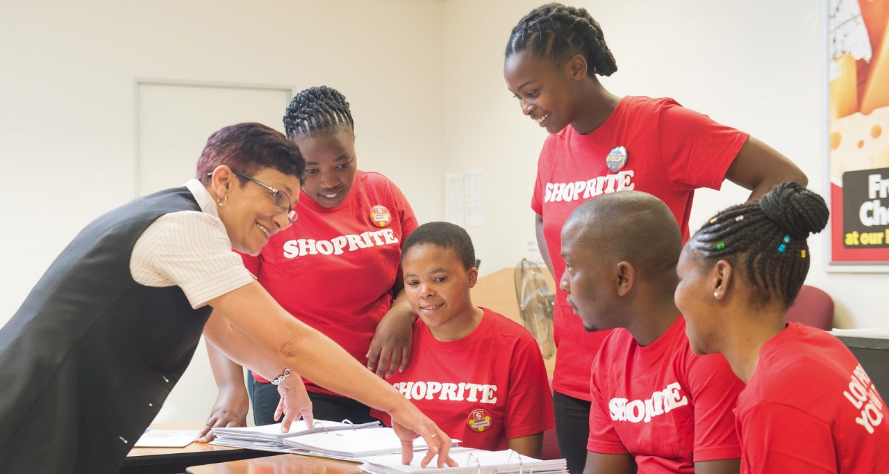 A group of students in red Shoprite t-shirts taking a lesson from one of the company employees. 