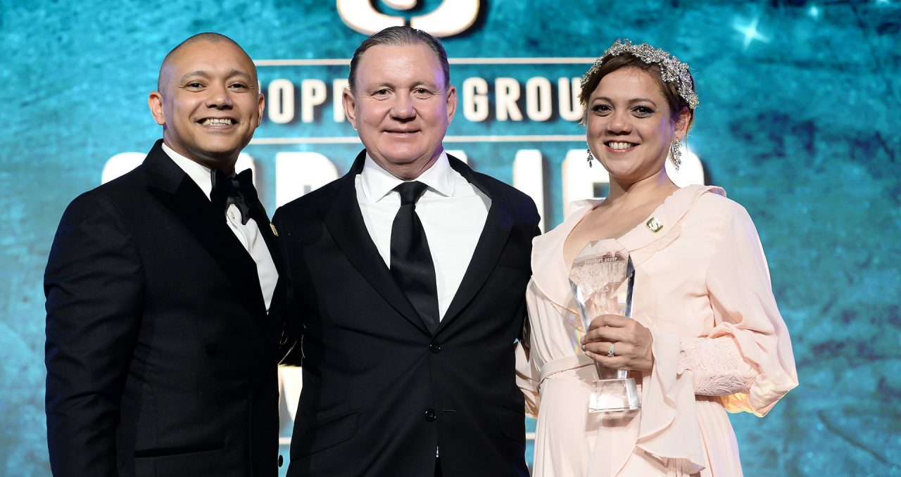Two gentleman in black suits and a lady on a cream colour dress holding an award.